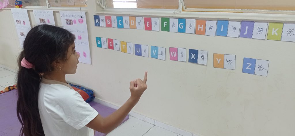 Uma menina de cabelos longos e amarrados faz o sinal da letra “i” em frente a um alfabeto em português e Libras disposto na parede da sala de aula. Fim da descrição.