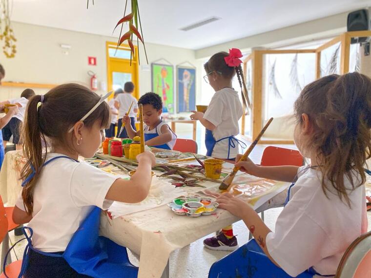 Descrição da imagem: Um grupo de crianças está em volta de uma mesa, a maioria sentada. Um em pé. Elas participam de uma atividade artística com tinta e pincel. Fim da descrição.