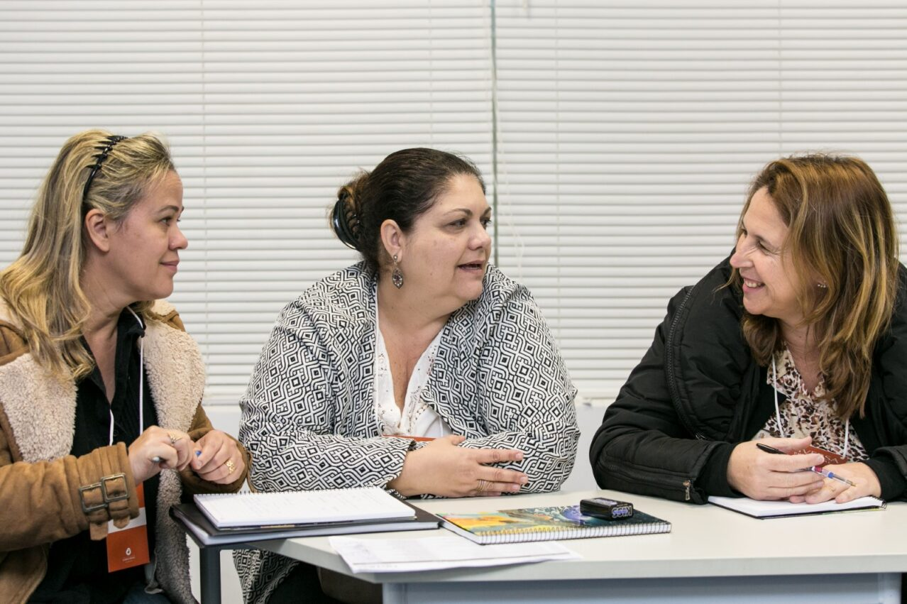 Três mulheres brancas conversam sentadas ao redor de uma mesa: uma loira, um cabelo castanho-escuro e a outra com mechas loiras. Duas estão com cadernos e canetas. Fim da descrição