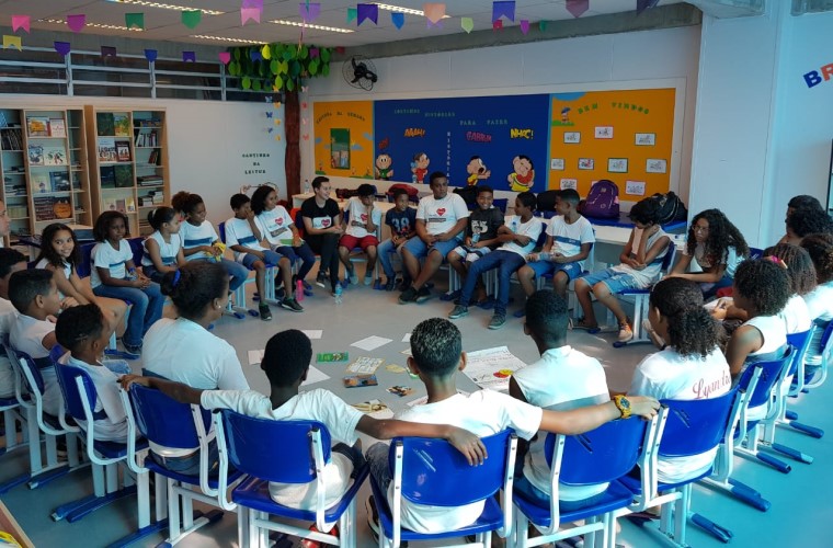 Em sala de aula, grupo de estudantes estão sentados em roda juntamente com jovens participantes do Rede Não Bate, Eduque durante momento de busca ativa. Fim da descrição.