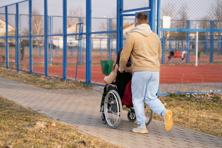 Em uma área externa, um homem de calça azul e moletom marrom claro empurra a cadeira de rodas de uma mulher. Ambos estão de costas, próximos a uma quadra de esportes. Fim da descrição.
