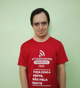 Homem com Síndrome de Down, branco, de cabelos curtos e escuros. Está usando camiseta vermelha com a frase Faça com a gente, não pela gente. Fim da descrição