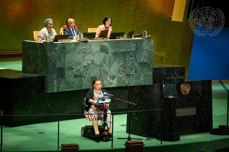 Na sala de conferências da Organização das Nações Unidas, a ONU, de Nova York, Heba Hagrass está no centro do palco, diante do microfone. Ela é uma mulher de pele branca, com cabelos curtos e castanhos e faz uso de uma cadeira de rodas motorizada. Atrás dela, na parte mais alta do palco, estão um homem e duas mulheres sentados. Fim da descrição.