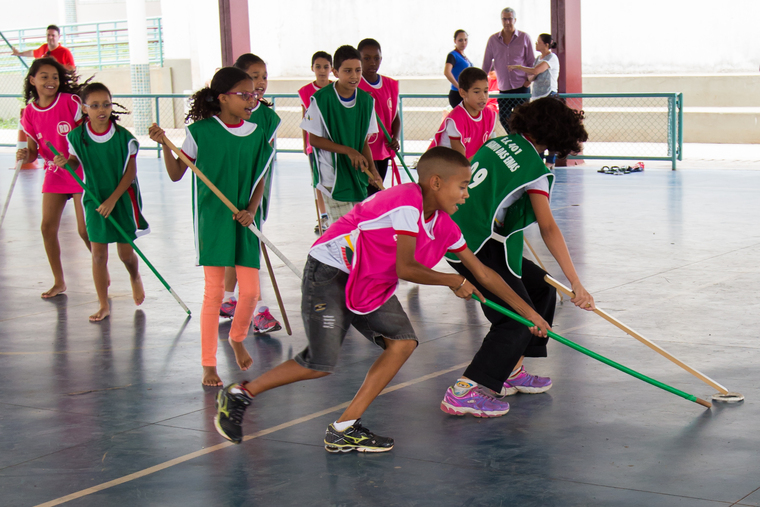 Grupo de crianças está em quadra escolar enquanto joga hockey. Elas estão divididas em dois times, no qual um usa colete rosa e outro verde. Todos seguram cabos de vassoura, utilizada para ser o taco do jogo. Em destaque, uma menina e um menino disputam a posse do disco. Fim da descrição.