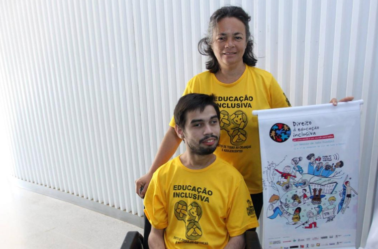 Na imagem, Keila Leite Chaves e Antonio Davi Sousa vestem camisetas amarelas com a frase "Educação Inclusiva" e ilustrações relacionadas ao tema. Davi, um jovem de pele clara e cabelos castanhos escuros, utiliza uma cadeira de rodas. Keila está em pé, atrás dele. Ela tem a pele clara, cabelos castanhos e mechas grisalhas. Ambos sorriem para a foto. Ela segura um banner com a frase "Direito à Educação Inclusiva" no topo. No centro do banner, uma ilustração colorida retratar diversas situações relacionadas à educação e ao cotidiano de pessoas com deficiência. Ambos sorrindo. O fundo da imagem mostra uma parede branca com ripas verticais. Fim da descrição.