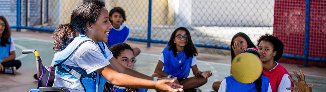 Uma jovem sorridente com cabelo escuro e encaracolado está sentada numa cadeira de rodas numa quadra polidesportiva ao ar livre. Ela está de perfil, virada para a direita, estendendo os braços para a frente como se tivesse acabado de lançar uma bola amarela. À sua volta, outras crianças e jovens estão sentados no chão da quadra formando um círculo, todos a usar coletes desportivos azuis sobre t-shirts brancas. No chão, no lado direito da imagem, uma jovem sentada acompanha o movimento da bola amarela em suspensão no ar. Ao fundo, vê-se uma rede metálica de proteção da quadra.