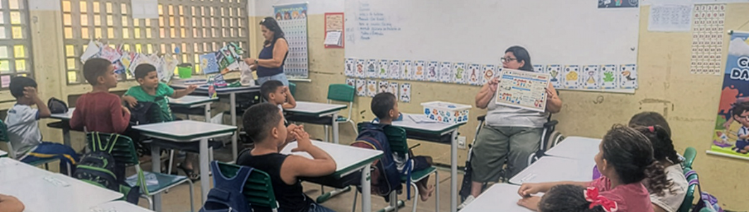 Sala de aula com estudantes sentados em carteiras, voltados para uma professora em cadeira de rodas, no centro da sala, segurando um material didático ilustrado. Mais ao lado, outra professora organiza materiais próximos à lousa, enquanto cartazes educativos e trabalhos dos estudantes decoram as paredes.