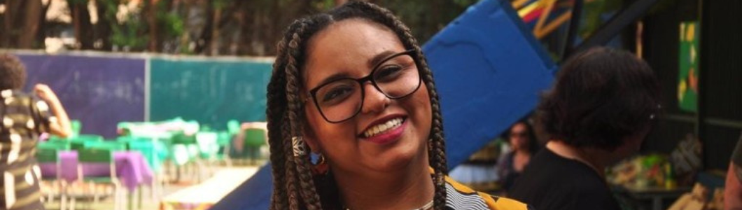 Amanda Santana, mulher negra, com tranças longas castanhas, óculos de armação preta e brincos, sorri para a câmera. Ela usa blusa amarela com estampa geométrica preta e branca. Ao fundo, há mesas com toalhas coloridas em um espaço ao ar livre.