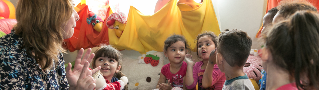 Professora sentada em roda com crianças pequenas em uma sala de educação infantil. Enquanto gesticula com as mãos, as crianças observam e interagem com ela. O ambiente é colorido, com almofadas, bonecos e brinquedos ao fundo.