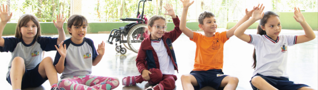 Cinco crianças sentadas no chão de uma quadra escolar participam de uma brincadeira coletiva com as mãos levantadas. Entre elas, há uma menina que faz uso de cadeira de rodas, reforçando a importância de propostas lúdicas que garantam a participação e a convivência entre todas as crianças no ambiente escolar.