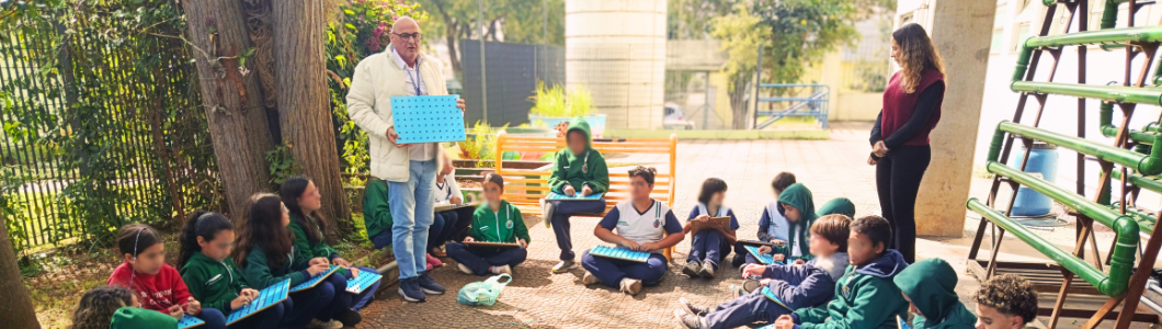 Grupo de estudantes participa de atividade ao ar livre conduzida por educadores. As crianças estão sentadas em círculo utilizando materiais didáticos durante uma proposta coletiva de aprendizagem.