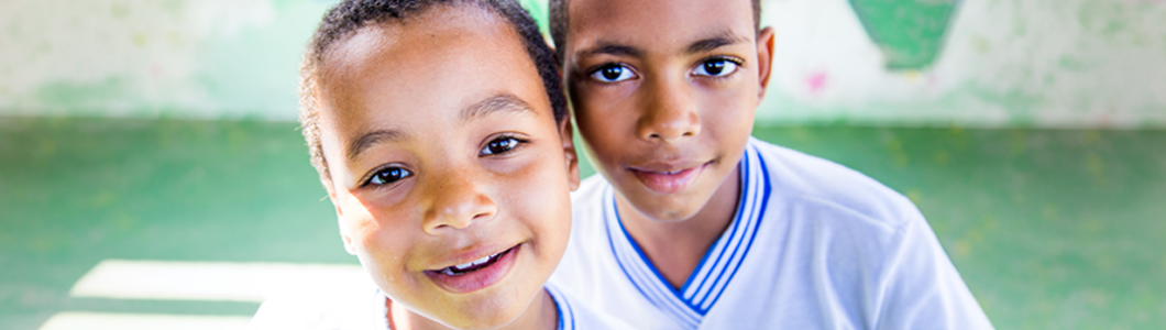 Fotografia em primeiro plano de duas crianças sorrindo, lado a lado, olhando diretamente para a câmera. Ambas vestem camisetas brancas com gola azul, semelhantes a uniformes escolares. Ao fundo, há um ambiente desfocado com tons de verde e elementos coloridos.