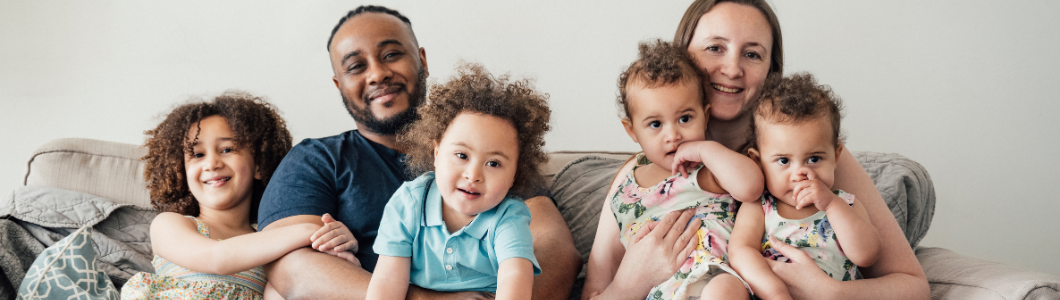 A imagem mostra uma família sentada em um sofá, em um ambiente interno claro e acolhedor. Há dois adultos e quatro crianças. À esquerda, uma criança mais velha, com cabelo cacheado, sorri levemente enquanto se inclina em direção ao grupo. Ao lado dela, um homem adulto está sentado com postura relaxada, sorrindo suavemente, com um dos braços apoiado atrás das crianças. À sua frente, uma criança pequena, também de cabelo cacheado, veste uma camisa azul clara e olha diretamente para a câmera. À direita do homem, uma mulher adulta segura duas crianças pequenas no colo, uma em cada braço. As duas crianças usam roupas claras com estampas florais e levam as mãos à boca, em gestos semelhantes. A mulher sorri para a câmera, transmitindo proximidade e cuidado. O fundo é neutro, com uma parede clara, e o sofá tem tons suaves, reforçando uma sensação de conforto, afeto e convivência familiar.