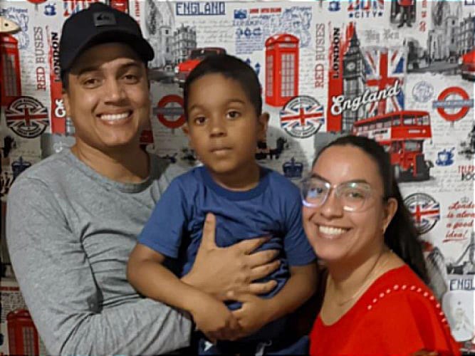 Foto de Waldilaine, ao lado do filho Dilan e o marido Denis, que posam para foto. À esquerda está Denis, um homem negro, com cabelos curtos e veste boné preto e camiseta cinza. Ele segura no colo o filho Denis, um menino negro, de cabelos escuros e curtos, que veste camiseta azul. À direita está Waldilaine, uma mulher branca, de cabelos escuros, longos e preso, que veste camiseta vermelha e óculos. Fim da descrição.