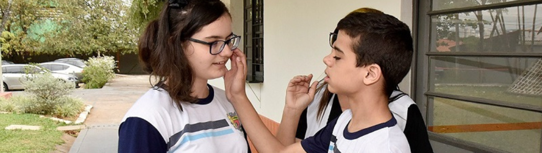 Fotografia de dois estudantes uniformizados conversando em Libras, em frente à escola. O estudante à direita movimenta as mãos próximo ao rosto da colega, que usa óculos e acompanha seus gestos.