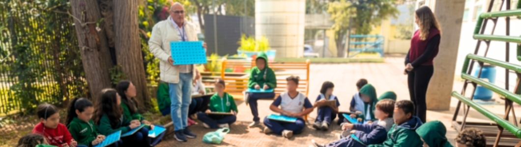 Grupo de estudantes participa de atividade ao ar livre conduzida por educadores. As crianças estão sentadas em círculo utilizando materiais didáticos durante uma proposta coletiva de aprendizagem.