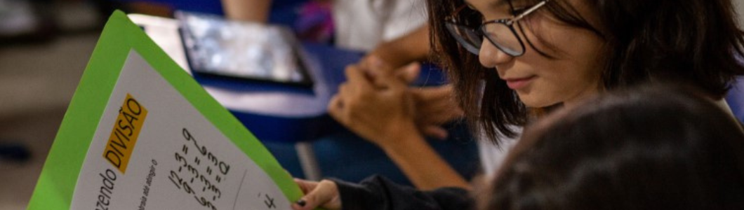 Estudantes em sala de aula. Em destaque, duas meninas, uma com máscara e outra sem, olham para o “caderno de cartazes”, que mostra maneiras de como solucionar operações matemáticas básicas. Fim da descrição.