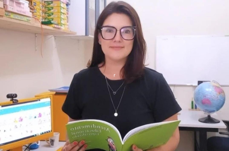 Foto de Nathalia Meneghine, uma mulher branca e de cabelos escuros na altura dos ombro. Ela veste uma blusa preta, óculos de grau e está com um livro de capa verde aberto nas mãos. Ao fundo, há um globo terrestre de mesa sobre uma mesa e do um monitor de computador. Fim da descrição.