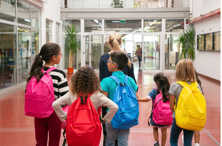 Um grupo de seis crianças andando pela escola junto a uma mulher adulta. Entre as crianças, há cinco meninas e um menino, todas com mochilas coloridas nas costas. A imagem mostra todas elas de costas. Fim da descrição.