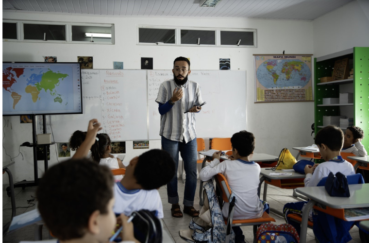 Foto do professor Leonardo Duarte Salomão, de 23 anos de idade, dentro de uma sala de aula falando com seus alunos. Ele é um homem de pele clara, tem cabelo e barba escuros e usa óculos de grau. Ele veste uma blusa azul de mangas compridas, uma camisa de listras por cima, uma calça jeans azul e calça sandálias. Fim da descrição.