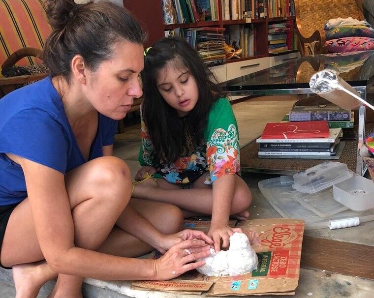 Maria Antônia e Beatriz estão sentadas no chão de uma sala de estar, lado a lado, fazendo uma massa com as mãos. Maria é uma mulher de pele branca e cabelo castanho, preso em um coque. Ela usa uma blusa azul e um short preto. Beatriz é uma criança de pele branca e cabelo castanho escuro. Ela usa um vestido verde florido. Fim da descrição.