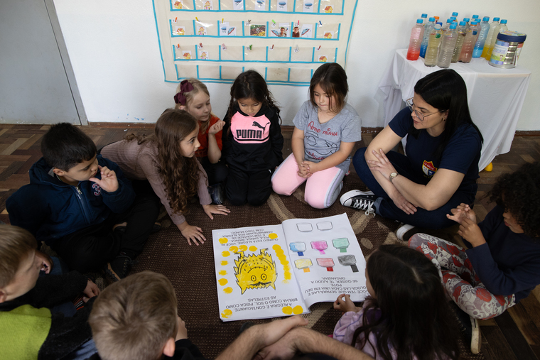 Em uma sala de aula de educação infantil, um grupo de nove crianças, sendo uma negra e as demais brancas, está reunido em torno do livro "O Monstro das Cores". Meninas e meninos de cinco anos estão sentados no chão, em roda, ao lado da professora, que é uma mulher branca de cabelos escuros. Fim da descrição.