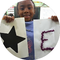 Menina segura dois cartazes, um com uma estrela preta e outro com a letra “E” em colagem.