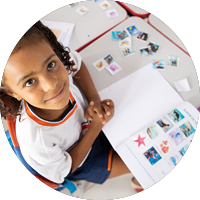 Menina de uniforme branco com detalhes laranja sorri para cima, com caderno aberto e figuras coloridas na mesa.