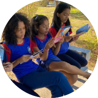 Três meninas de uniforme azul e vermelho leem livros sentadas em banco ao ar livre.
