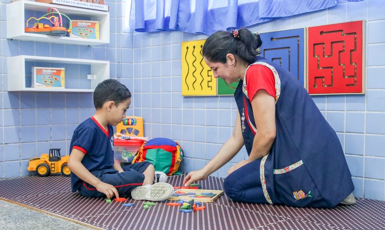 Em uma sala com brinquedos, um menino branco com uniforme está sentado no chão enquanto brinca de peças de montar com sua professora, uma mulher branca que está agachada ao seu lado. Fim da descrição.