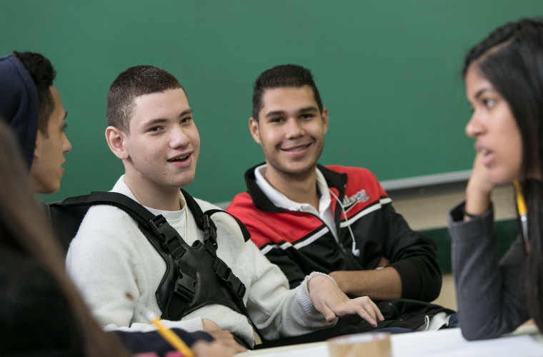 Grupo de estudantes do Ensino Médio sentados em uma sala de aula, organizados em círculo, em momento de conversa e interação. No centro da imagem, um estudante com deficiência física utiliza um colete de apoio no tronco e participa ativamente da conversa, com expressão atenta e gestos com as mãos. Ao seu lado, outro estudante sorri enquanto observa o diálogo. À direita, uma estudante aparece de perfil, com a mão apoiada no rosto, escutando atentamente. Ao fundo, há um quadro verde típico de sala de aula. A cena transmite troca, participação e convivência entre os estudantes em um ambiente escolar inclusivo. Fim da descrição.