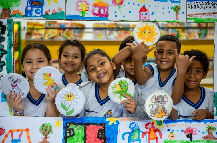 Seis crianças em sala de aula sorriem enquanto seguram desenhos pintados em círculos de papel, como sol, flor, sapo e cachorro. Elas estão posicionadas atrás de um painel decorado com desenhos colorido.