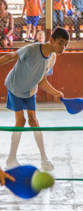 Estudante em quadra escolar jogando com raquete azul e bola verde sobre rede baixa, enquanto outras crianças observam ao fundo, atrás de uma grade.