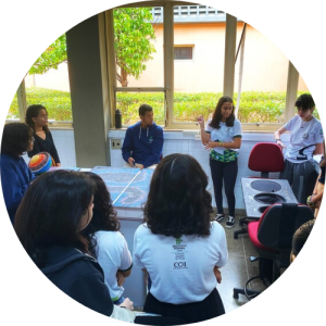 Grupo de estudantes do Ensino Médio reunido em um laboratório escolar, em pé e em círculo, durante uma atividade coletiva. No centro, uma estudante fala ao grupo enquanto segura materiais didáticos, indicando um momento de explicação ou apresentação. Outros estudantes observam atentamente, alguns com mochilas e roupas escolares. O ambiente conta com bancadas de laboratório, cadeiras giratórias e equipamentos sobre as mesas. Ao fundo, grandes janelas deixam entrar luz natural e revelam árvores e áreas verdes do lado de fora da escola. A cena expressa participação, escuta e aprendizagem colaborativa em um espaço educativo. Fim da descrição.