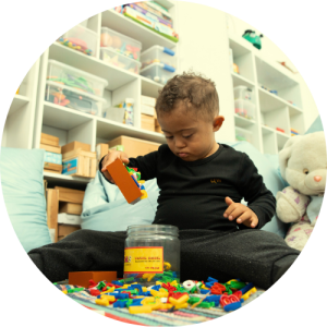 Criança pequena sentada no chão de uma sala com prateleiras de brinquedos, brincando com peças de montar coloridas ao lado de um pote transparente.