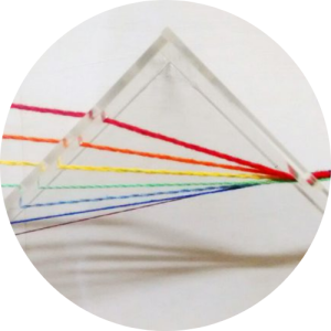 Três placas de acrílico formam um triângulo. A luz branca, representada pela união de diferentes fios de crochê, atravessa uma das placas e, dentro do prisma, se dispersa em sete fios de cores e texturas diferentes. Esses fios saem atravessando outra placa de acrílico. Fim da descrição. 