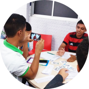 Em sala de aula e ao redor de mesa escolar, quatro estudantes interagem com o jogo da argumentação. Um deles grava com o celular fala de colega. Fim da descrição.