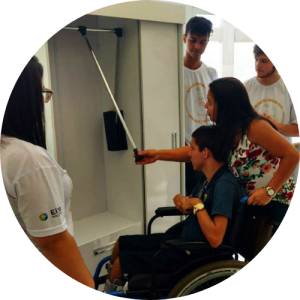 Jovem com deficiência física observa guarda-roupa adaptado juntamente a mulher que segura sua cadeira de rodas. Ao lado, três estudantes assistem. Fim da descrição.