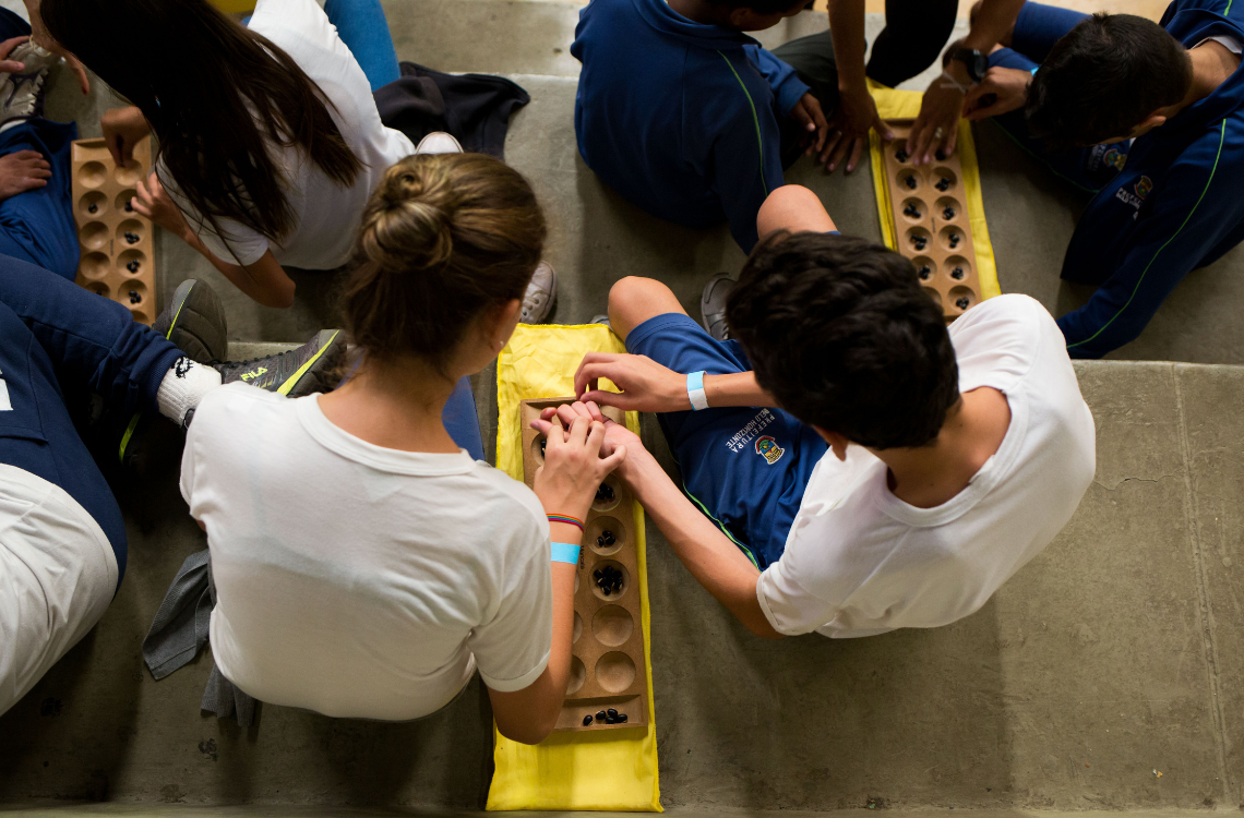 Vista superior de um grupo de crianças sentadas no chão, em ambiente escolar, realizando juntas uma atividade pedagógica com um tabuleiro de madeira contendo cavidades e peças pequenas. As crianças usam uniforme escolar e manipulam o material de forma colaborativa, com apoio de um adulto.