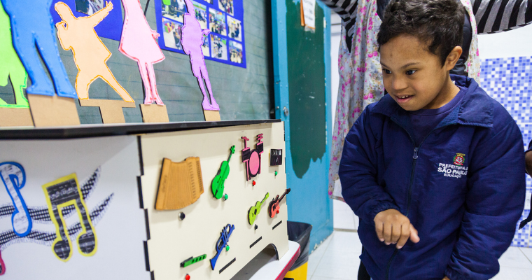 Um menino com síndrome de Down está em um ambiente escolar, posicionada à direita da imagem, sorrindo e interagindo com um painel pedagógico interativo. Ele veste um casaco azul com o logotipo “Prefeitura de São Paulo – Educação”. À sua frente, há um móvel claro com diferentes elementos coloridos fixados, como instrumentos musicais, números, símbolos e peças em relevo, pensados para estimulação sensorial e aprendizagem lúdica. Na parte superior do móvel, aparecem silhuetas de pessoas em cores vibrantes, recortadas e apoiadas em uma base. Ao fundo, vê-se uma parede azul com murais e fotos expostas, além de uma porta verde. Fim da descrição.
