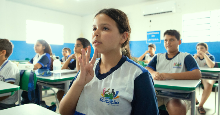 Em uma sala de aula do Ensino Fundamental, uma estudante com pele clara, cabelos castanhos presos e uniforme escolar levanta a mão enquanto acompanha a aula. Ela está sentada em uma carteira individual, com expressão atenta e concentrada. Ao fundo, outros estudantes, também uniformizados, permanecem sentados e atentos à atividade. O ambiente é bem iluminado, com paredes em tons de azul e branco, carteiras organizadas em fileiras e uma lousa visível, indicando o contexto de uma aula em escola pública.