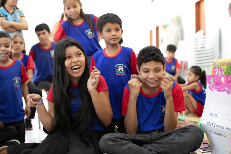 Fotografia de um grupo de crianças em uma sala de aula, todas vestindo um uniforme escolar composto por uma camiseta azul com mangas vermelhas e um brasão no peito. Em primeiro plano, uma menina de longos cabelos pretos sorri com entusiasmo, enquanto um menino ao seu lado, sentado de pernas cruzadas, olha para baixo com uma expressão de alegria contida, segurando um pequeno objeto nas mãos. Ao fundo, outras crianças observam a cena. Fim da descrição.