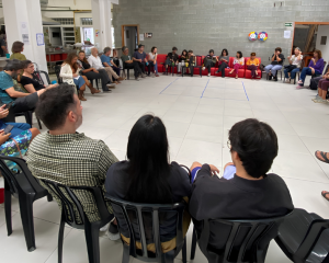 A foto mostra uma sala com diversos educadores reunidos em roda, sentados em cadeiras de plástico. Fim da descrição. Gestão