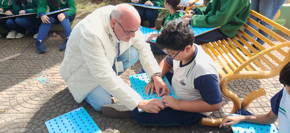 Professor auxilia estudante durante atividade realizada em espaço externo da escola. Ao redor, outros alunos participam da mesma proposta utilizando materiais pedagógicos adaptados. trabalho colaborativo