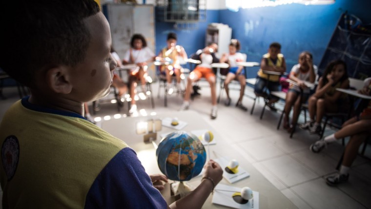 Criança em primeiro plano segura um globo terrestre durante atividade em sala de aula, enquanto outros estudantes estão sentados em círculo ao fundo.