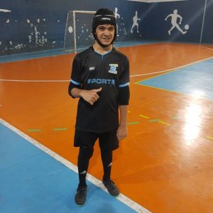 Jovem de cabelos escuros está em pé em uma quadra de futsal, usando uniforme preto, calça esportiva, tênis e uma proteção acolchoada preta na cabeça. Ele faz sinal de positivo com uma das mãos. Ao fundo, há uma trave e desenhos de jogadores de futebol na parede.