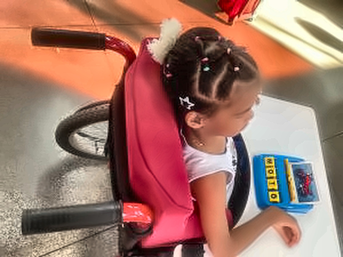 Menina sentada em uma cadeira de rodas, com o corpo voltado para uma mesa branca, onde há um recurso pedagógico colorido que ela formou a palavra "moto". Ela tem os cabelos presos em tranças decoradas com presilhas e veste uma blusa branca.
