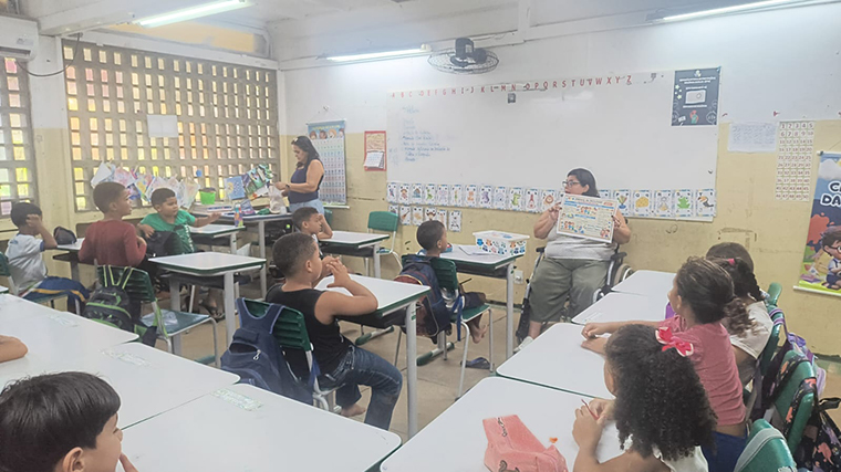 Sala de aula com estudantes sentados em carteiras, voltados para uma professora em cadeira de rodas, no centro da sala, segurando um material didático ilustrado. Mais ao lado, outra professora organiza materiais próximos à lousa, enquanto cartazes educativos e trabalhos dos estudantes decoram as paredes. Fim da descrição.