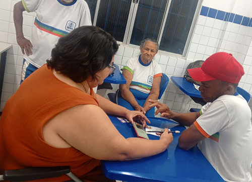 A foto mostra a professora Adriana Cunha, que é uma mulher branca de cabelos escuros vestindo uma blusa laranja, dando aula para alunos da Educação de Jovens e Adultos (EJA). Ela está sentada à mesa com um aluno, utilizando materiais didáticos, enquanto outros dois estudantes acompanham a atividade. O ambiente possui mesas e cadeiras escolares azuis e paredes revestidas de azulejos brancos. Fim da descrição.