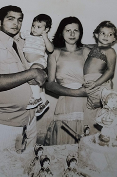 Fotografia em preto e branco de um casal com duas crianças pequenas, posando juntos em frente a uma parede clara. O homem segura uma das crianças no colo, enquanto a mulher segura a outra. À frente deles, há uma mesa com bonecas e outros brinquedos. Fim da descrição.